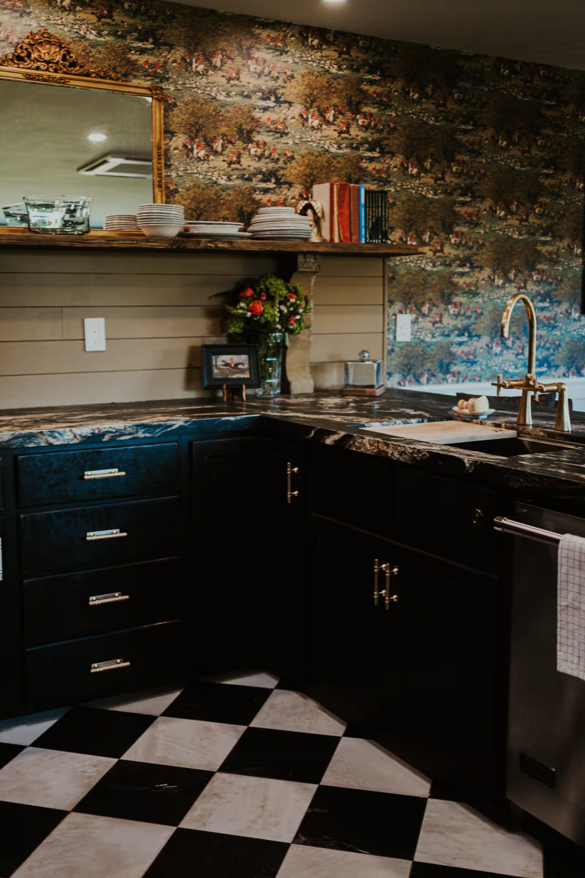 The kitchen with hunt-scene wallpaper, black cabinetry, marble counters, a brass gooseneck faucet, and a black-and-cream checkerboard floor