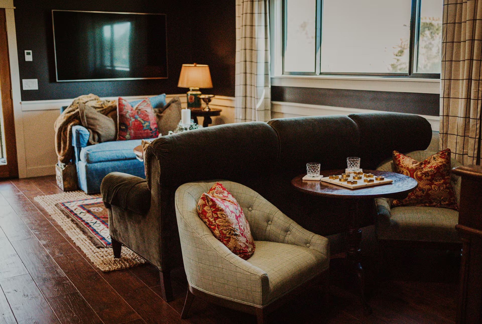 The lounge with a velvet chesterfield sofa, tufted plaid chairs, a checkers set, and a blue velvet loveseat beneath a wall-mounted TV