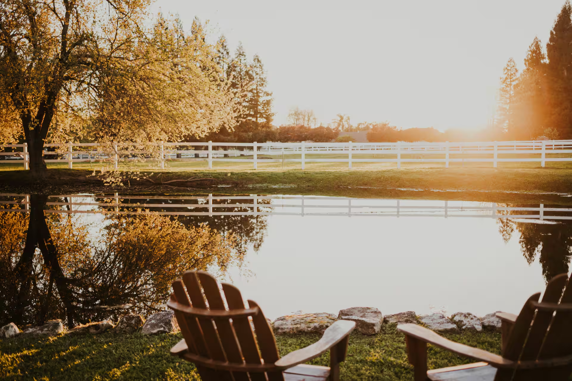 Two wooden Adirondack chairs on the grassy bank of the farm pond at golden hour, facing still water that mirrors the white horse fence and flowering trees